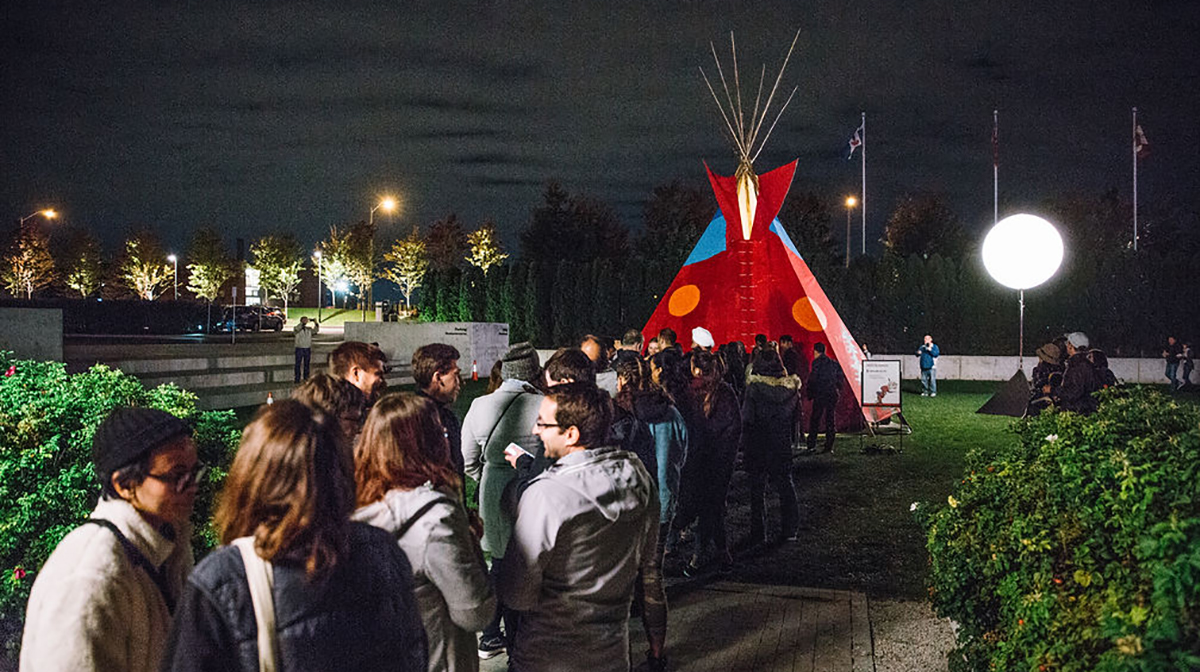 A lineup of happy people stand in line to get into a red teepee.